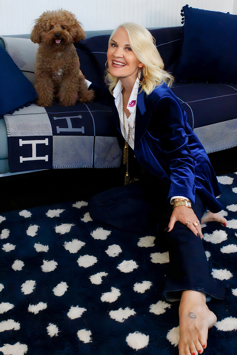 Jacqui Stafford in a blue velvet jacket sitting on a patterned rug with her dog seated on a couch.