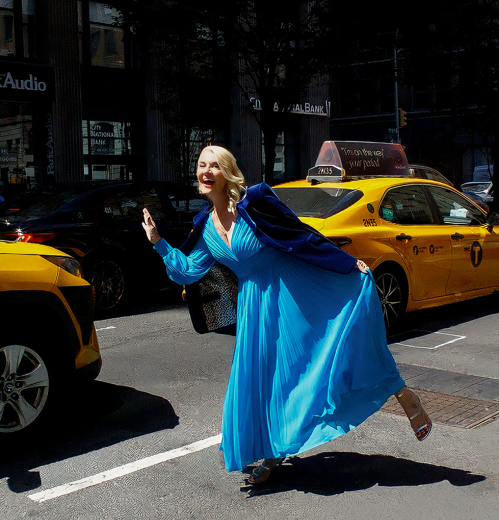 Jacqui Stafford pictured in a light blue dress crossing a busy New York CIty street with bright yellow taxis in the background.