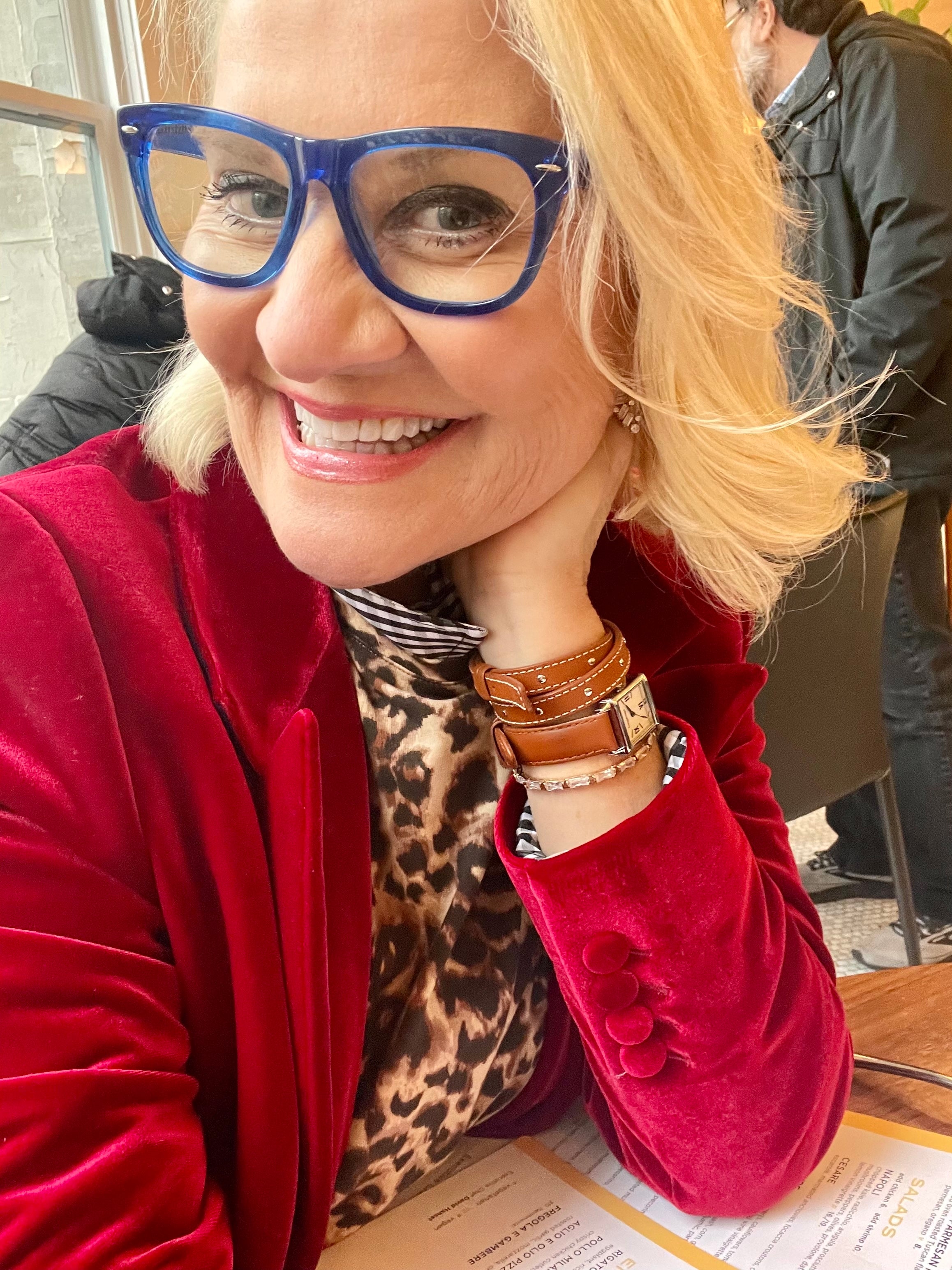 Jacqui Stafford fashion expert personal stylist portrait, blonde woman wearing stylish blue glasses, red velvet jacket, and leopard print top with leather watch